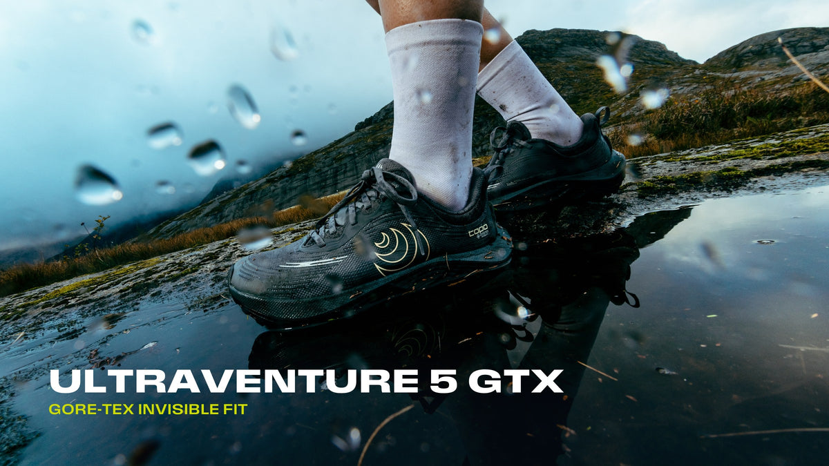 Topo Athletic Ultraventure 5 GTX i en vattenpöl på en berghäll, i regn. Text: Ultraventure 5 GTX, GORE-TEX Invisible Fit